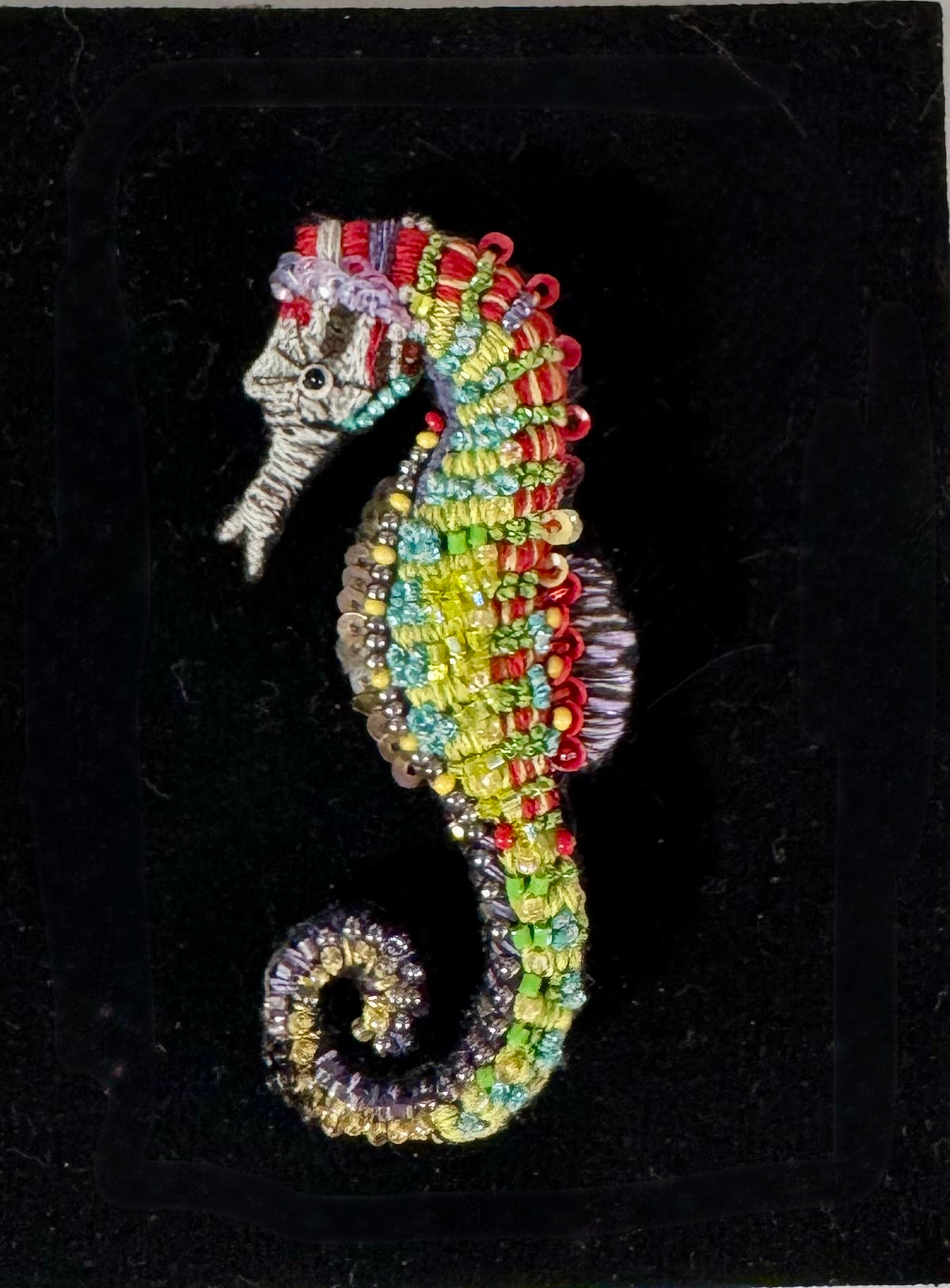 A colorful beaded seahorse brooch displayed on a black velvet background. The brooch features intricate beadwork in shades of green, yellow, red, blue, and purple, with silver-toned metal detailing on the head.