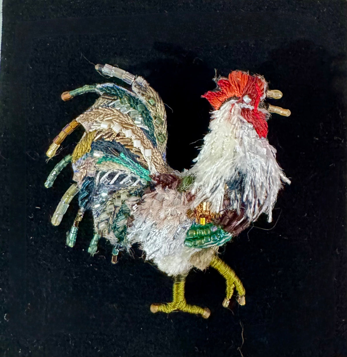 A handmade embroidered rooster figurine made from textured thread and yarn, featuring a red comb, white and gray body, and green and yellow accents, set against a solid black background.