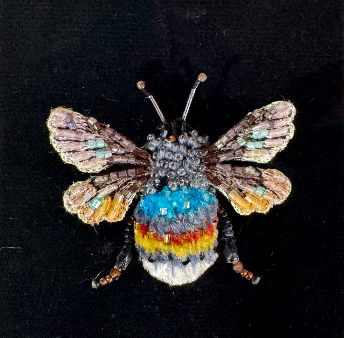 A detailed, handcrafted beaded bumblebee pin on a solid black background. The bee features intricate beadwork in shades of brown, beige, light blue, orange, yellow, red, and white, with metallic gold-toned antennae.